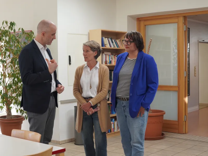 Ein Mann und zwei Frauen unterhalten sich in einem Raum, vorn ein Tisch, hinter ihnen eine Pflanze und ein B&uuml;cherregal.