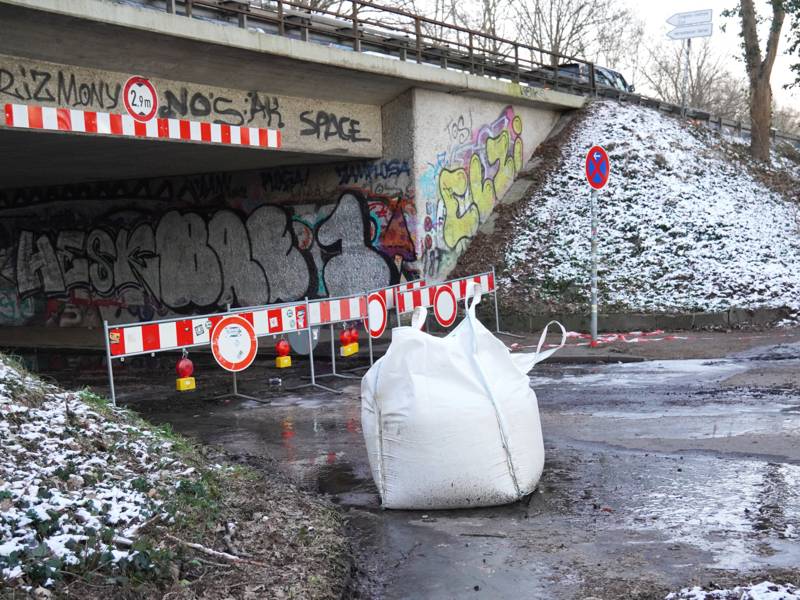 Ein weißer großer Sack, links dahinter rot-weiß gestreifte Schilder vor einer Unterführung, darüber eine Straße, Bäume auf der Böschung, leicht mit Schnee bedeckt.