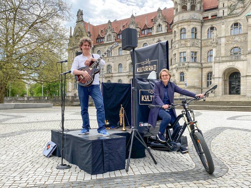 Ein Mann steht auf einer kleinen Bühne, die von einem umgebauten Fahrrad gezogen wird; auf dem Rad sitzt eine Frau. Im Hintergrund das Neue Rathaus in Hannover.