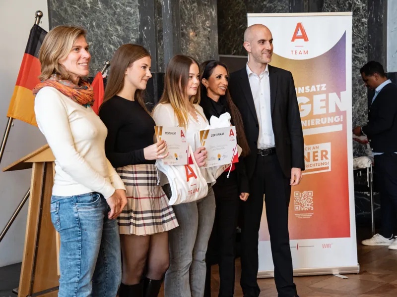 Drei junge Frauen halten ein Zertifikat und ein Shirt mit der Aufschrift A-Team in den H&auml;nden, links steht eine Frau, rechts ein Mann von einem Banner mit der Aufschrift A-Team.