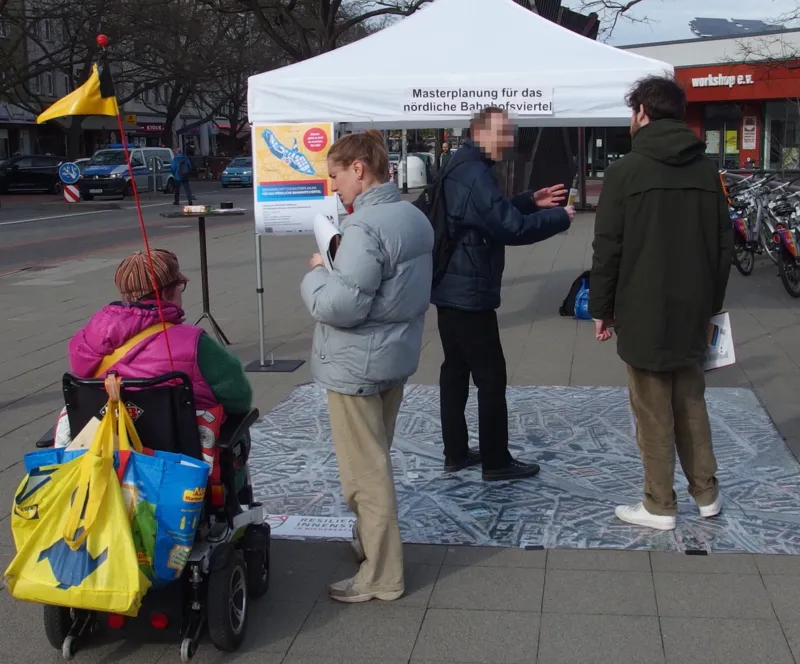 Auf dem Bild sind vier Personen zu sehen. Ganz links sitzt eine Person in einem elektrischen Rollstuhl und unterh&auml;lt sich mit einer rechts daneben stehenden Person, die einen Notizblock in der Hand h&auml;lt. Etwas im Hintergrund stehen zwei weitere Menschen, das Gesicht des einen ist unkenntlich gemacht. Auch sie unterhalten sich miteinander. Weiter im Hintergrund ist ein Pavillon zu sehen, der die Aufschrift "Masterplanung f&uuml;r das n&ouml;rdliche Bahnhofsviertel" tr&auml;gt.