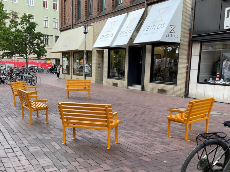 F&uuml;nf gelbe B&auml;nke stehen im Halbkreis auf einem geplastertem Gehweg vor Gesch&auml;ften.