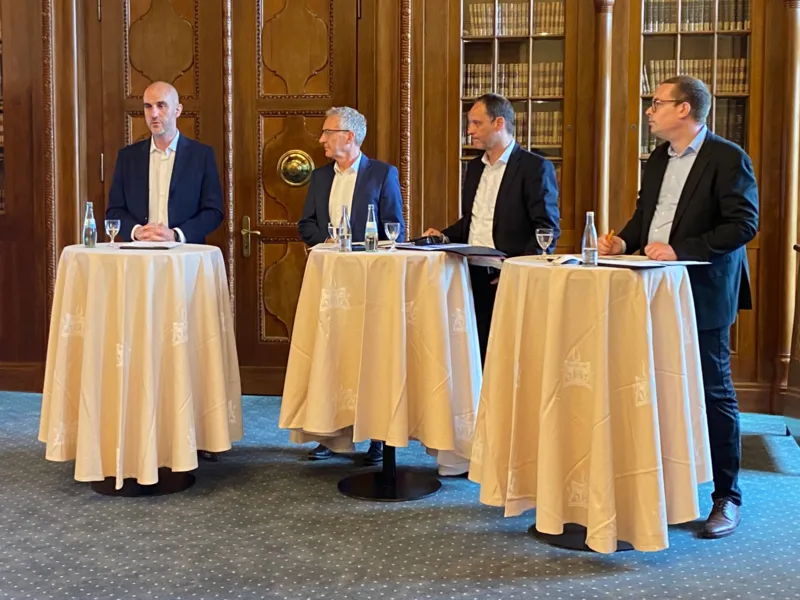 Vier M&auml;nner stehen je vor drei Stehtischen, einer auf der linken Seite spricht.