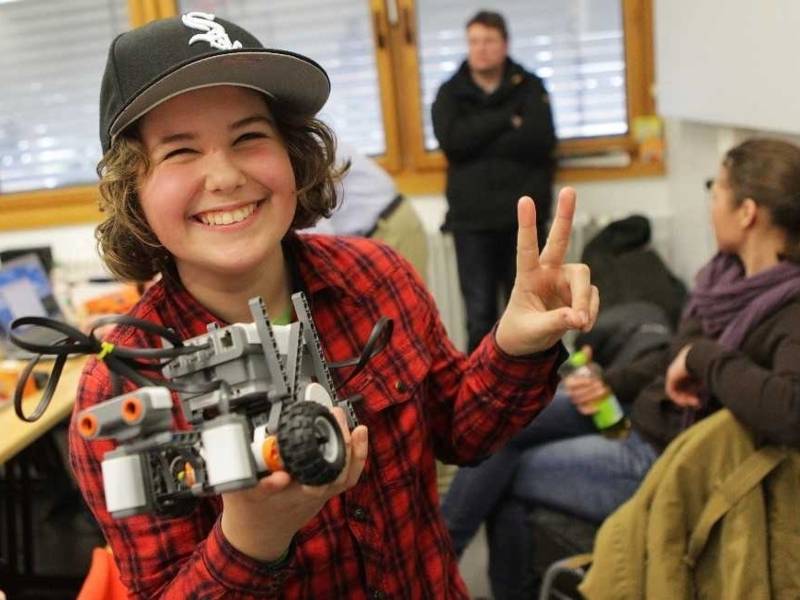 Lachendes M&auml;dchen mit Basecap h&auml;lt eine kleine Roboterkonstruktion und macht mit der anderen Hand das Victory-Zeichen.