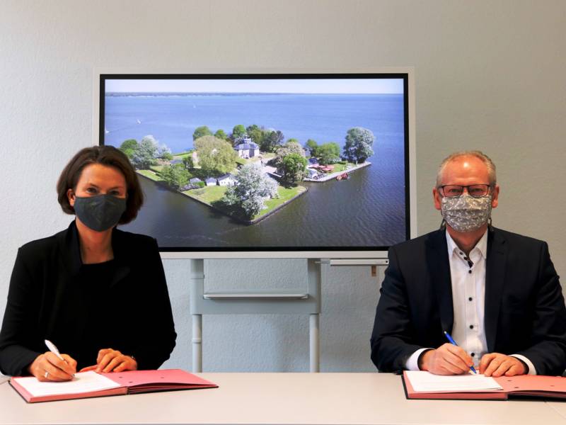 Zwei Personen bei einer Vertragsunterzeichnung. Im Hintergrund zeigt ein Bildschirm eine Inselfestung - die Insel Wilhelmstein im Steinhuder Meer