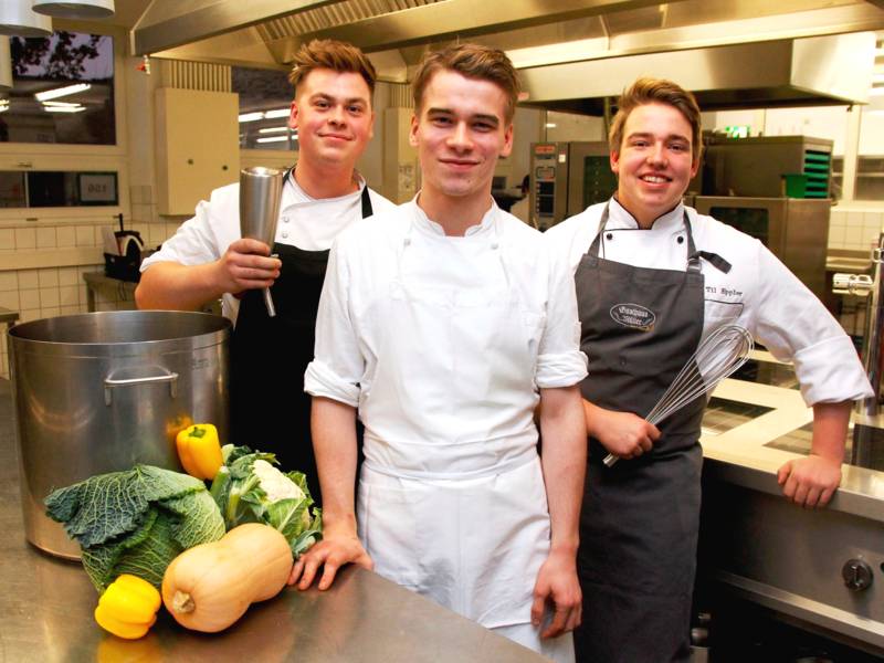 Drei Männer tragen die Arbeitskleidung eines Kochs und stehen in einer Großküche zwischen Lebensmitteln und Kochutensilien.