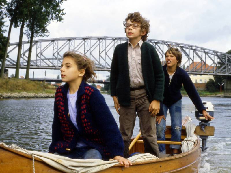Drei Kinder auf einem kleinen Motorboot auf einem Fluss, im Hintergrund eine Brücke.