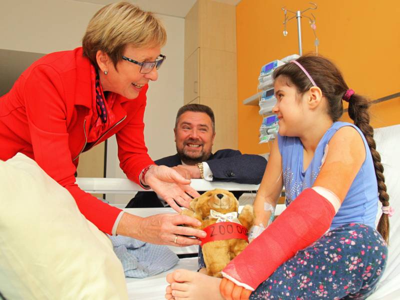 Ein Mädchen im Krankenhausbett freut sich über einen Teddybären, der ihr von einer Frau und einem Mann überreicht wird.