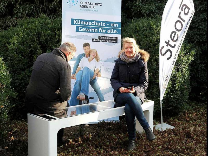 Ein Mann und eine Frau sitzen auf einer Parkbank vor einem Plakat mit der Aufschrift "Klimaschutz - ein Gewin für alle."