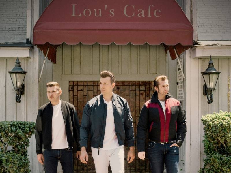 Drei Männer mit hochgestyleten Haaren und Bomberjacken an, stehen vor einem Café.
