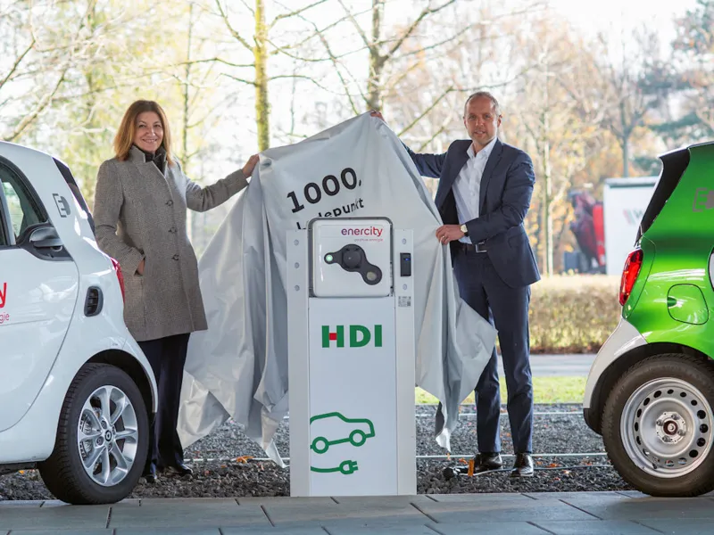 Eine Frau und ein Mann zwischen zwei Autos neben einer E-Lades&auml;ule heben eine Abdeckung hoch