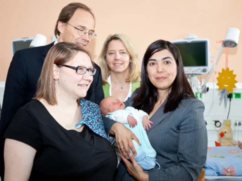 Drei Frauen und ein Mann stehen zusammen mit einem Neugeborenen in einem Krankenhauszimmer.