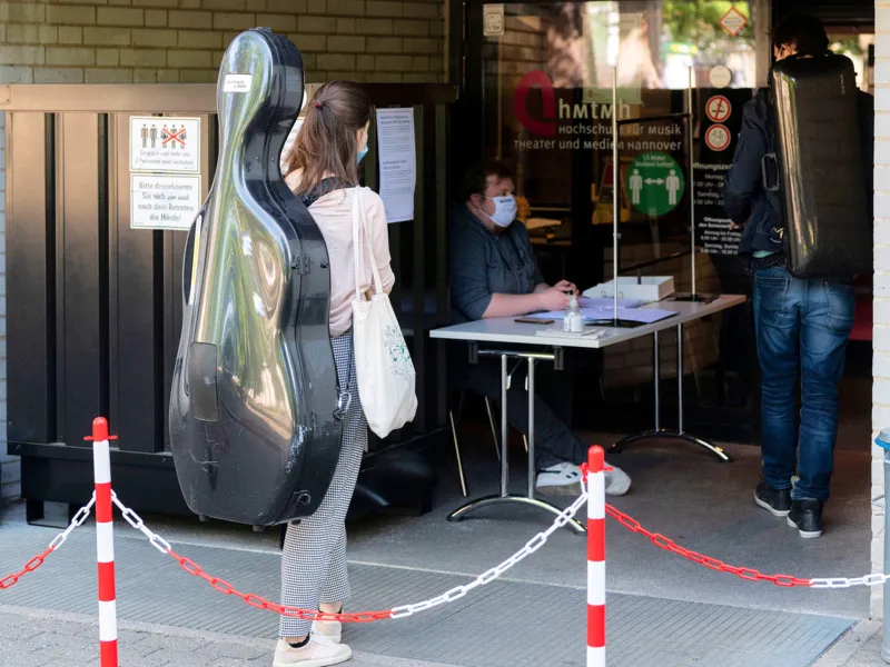 Frau mit Nasen- und Mundschutzmaske und Musikinstrumentenkoffer auf dem R&uuml;cken steht an einem kontrolliertem Eingangsbereich.