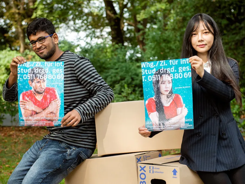Eine Frau und ein Mann halten jeweils ein Plakat mit frierenden Menschen im Regen und der Aufschrift "Lasst uns nicht im Regen stehen".