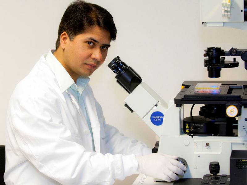 Mann in weißem Kittel mit weißen Handschuhen vor Mikroskop.