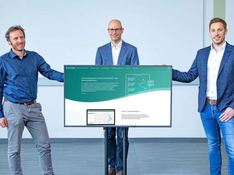 Drei M&auml;nner stehen neben und hinter einem Flachbildschirm.