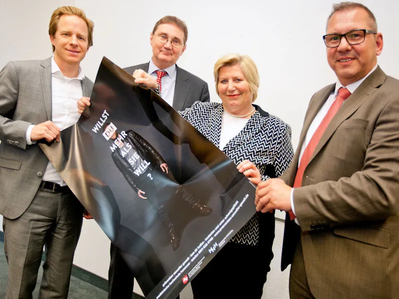 Eine Frau und drei M&auml;nner halten ein Plakat in den H&auml;nden mit der Aufschrift "Willst Du mehr als sie will?".