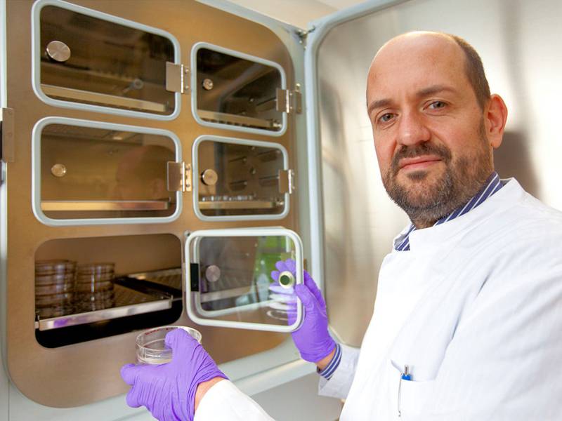 Mann in weißem Kittel vor geöffnetem Schrank mit Glastüren mit einer Petrischale in der Hand.