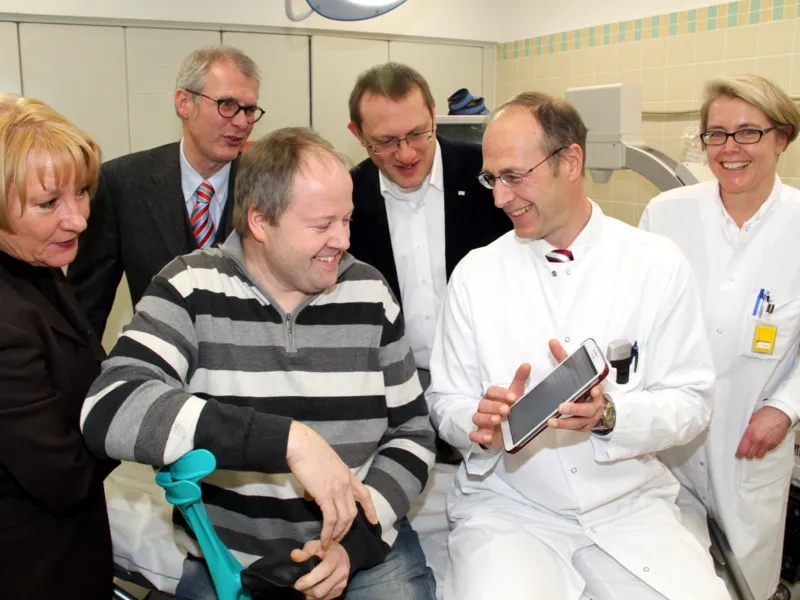 Ein Arzt im Kittel zeigt einem Mann mit Kr&uuml;cke ein Programm auf einem Tablet-Computer, drei weitere Personen sehen zu.