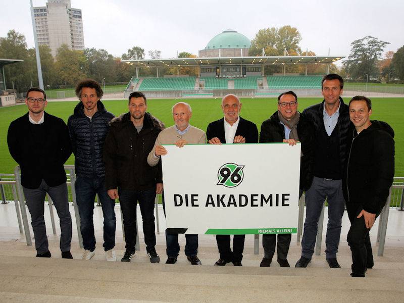 Gruppenbild mit acht Männern in einem Fußballstadion. 