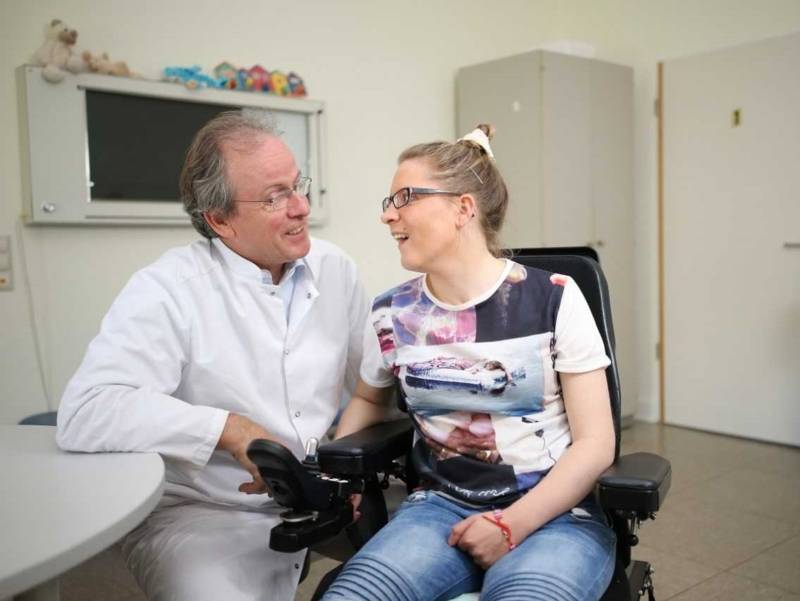 Klinikraum mit einem Arzt im Gespräch mit einer Patientin im Rollstuhl