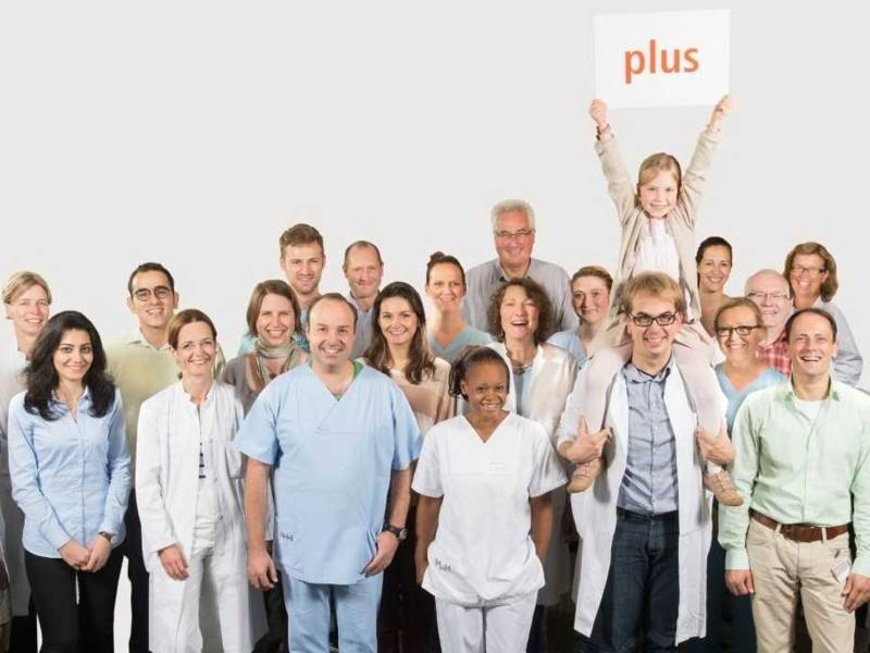 20 Frauen und Männer, teilweise in Arztkitteln, und ein Kind mit einem Schild mit dem Wort plus