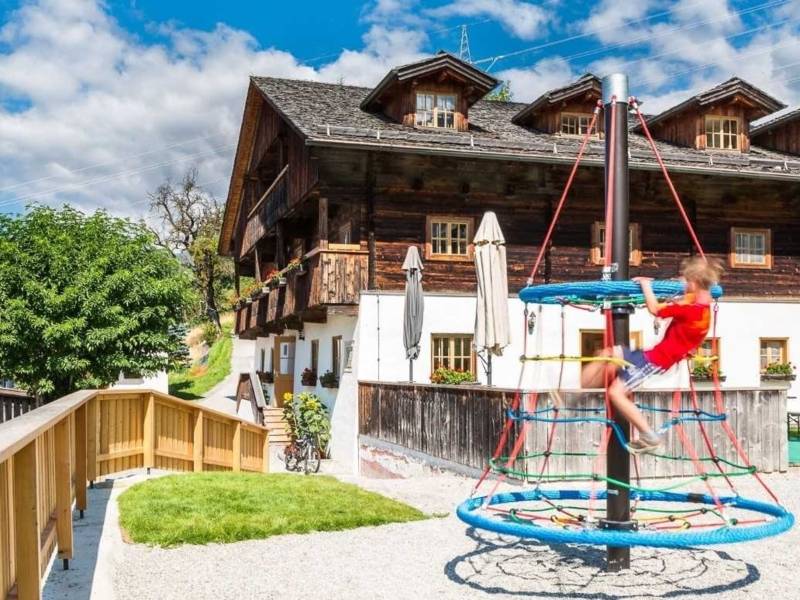 Großes Haus im alpinen Stil, davor ein Kind auf einem Klettergerät