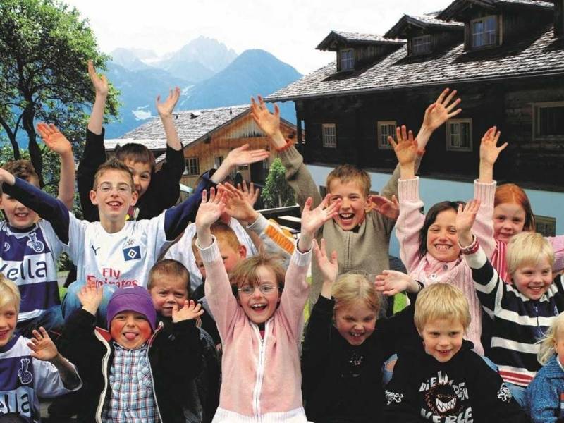 Gruppe von lachenden und winkenden Kindern, im Hintergrund ein Haus und Berge