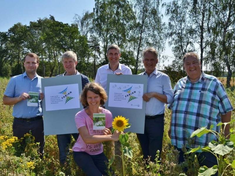 Eine Frau und f&uuml;nf M&auml;nner mit Bildern und Plakaten auf einer Wiese mit hohen Pflanzen