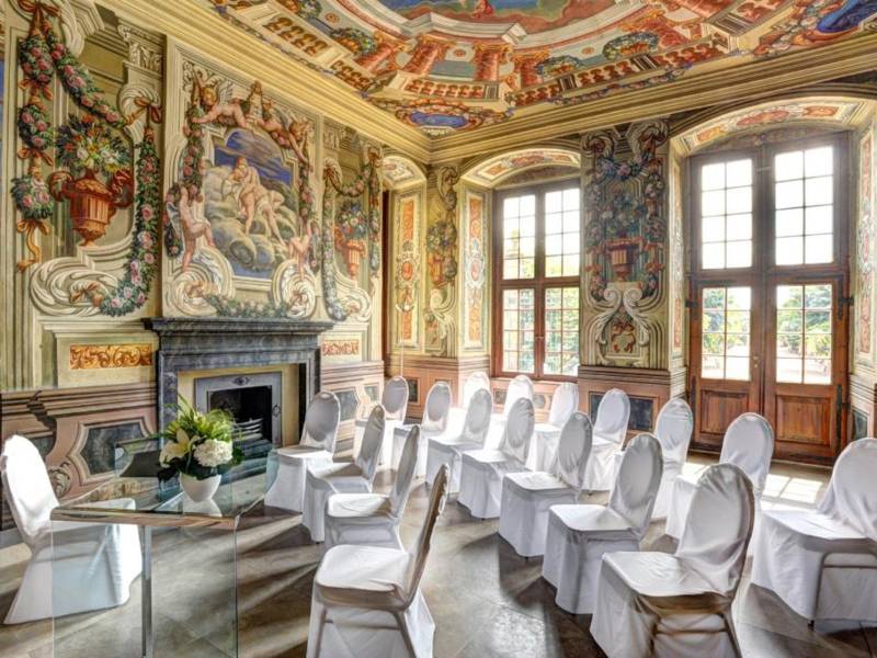 In einem mit zahlreichen Wandmalereien versehenen Raum mit hohen Fenstern sind vor einem Glastisch mit Blumenvase drei Stuhlreihen für eine Trauung aufgebaut.