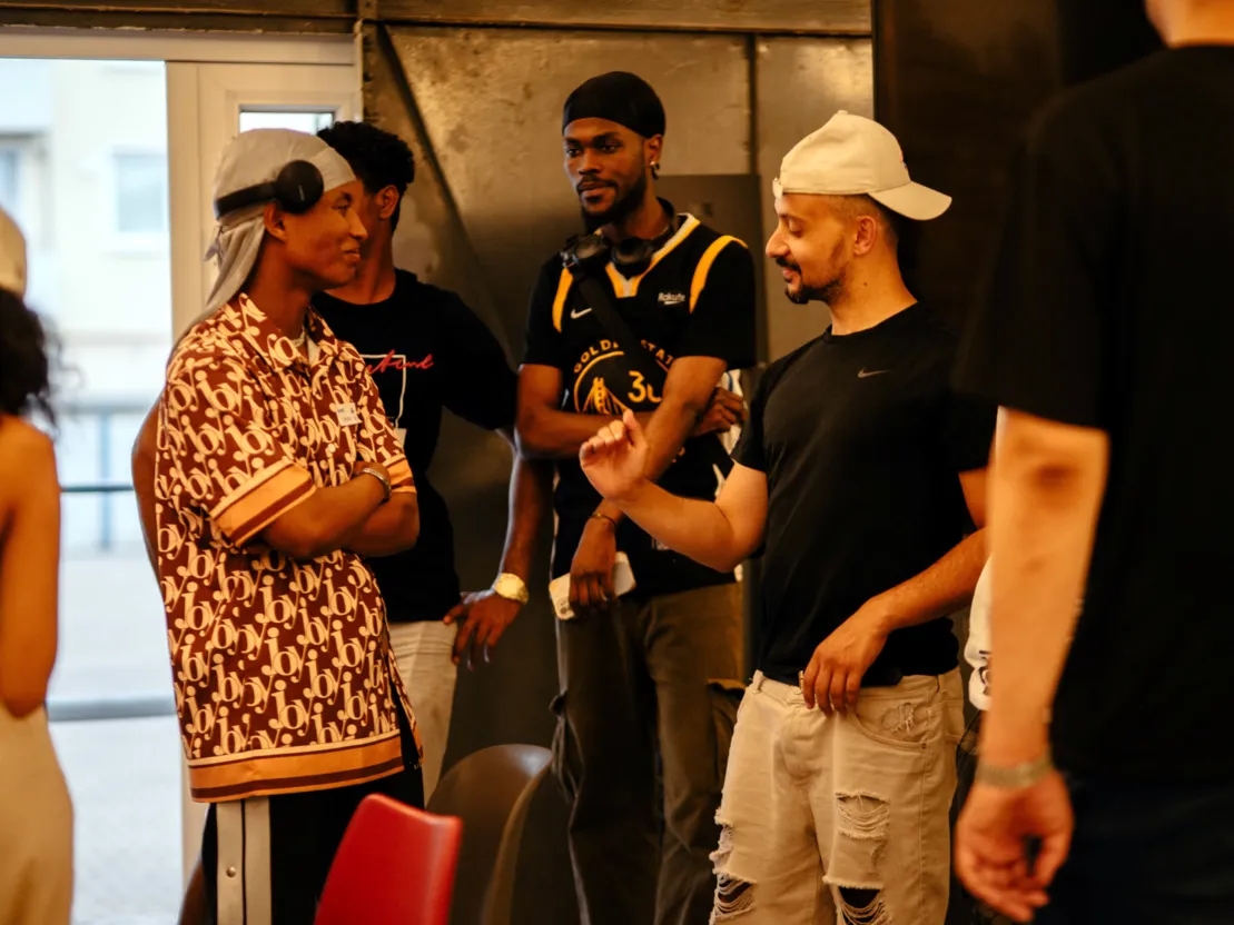 Vier Personen in der Bildmitte. Die Person rechts hat eine wei&szlig;e Basecap auf und scheint den anderen Personen etwas zu erkl&auml;ren.