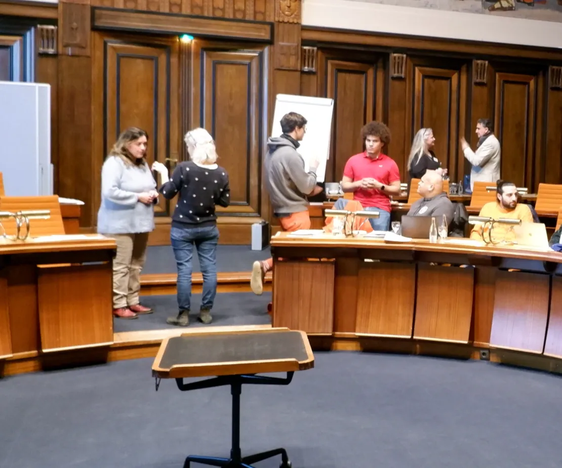 Acht Personen, teils stehend, teils sitzend, reden in kleinen Gruppen miteinander. Sie befinden sich in einem holzvert&auml;felten Raum mit halbrunden Plenartischen.