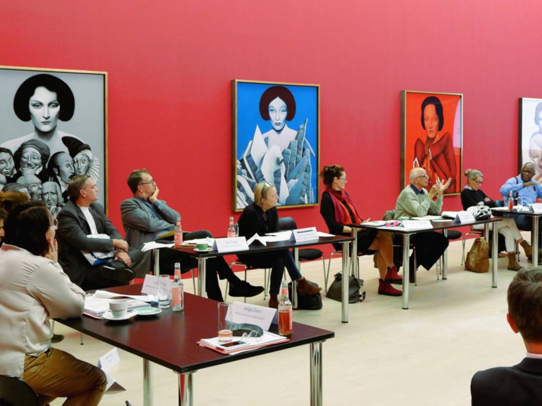 Elf Personen (5 Frauen und 3 Männer) sitzen in einem rot gestrichenen Raum an Tischen und schauen zur rechten Bildseite. Dort scheint ein Mann einen Redebeitrag zu halten. Auf den Tischen sind Arbeitsmaterialien, Namensschilder und Getränke zu sehen.