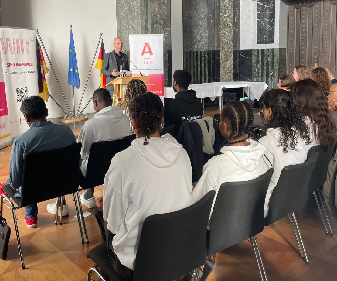 OB Onay spricht im Mosaiksaal vor jungen Menschen. Neben ihm stehen zwei Banner: Eines von den A-Teams, eines vom WIR2.0-Prozess.