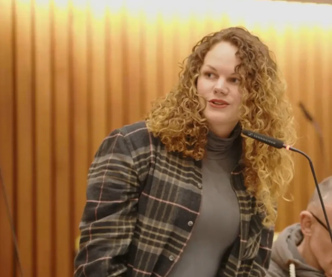 Eine Frau mit langen blonden Locken spricht in ein Mikrofon. Sie tr&auml;gt ein kariertes Hemd.