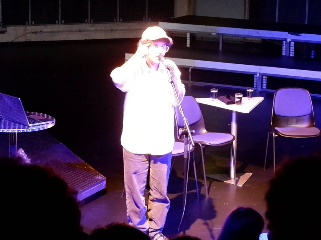 Eine Person mit lockigen Haaren, weißer Basecap und weißem Hemd steht am Mikrofon und singt. Der Bildhintergrund ist dunkel.