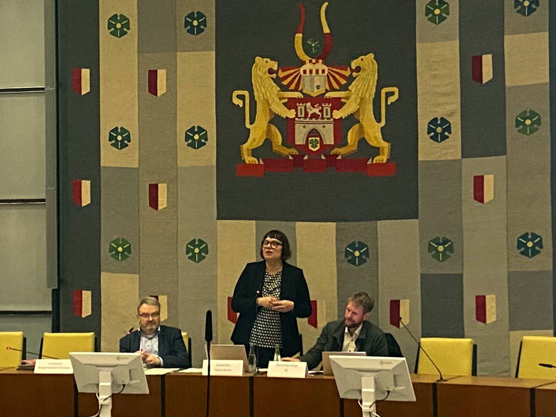 Drei Personen an einem gebogenen Plenartisch. Die Frau in der Mitte steht und spricht in ein Mikrofon, die Männer links und rechts von ihr sitzen.