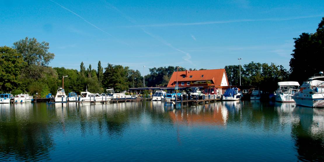 Wasserfläche mit Blick auf einen Yachthafen. Der Hafen wird durch Baumbestand eingerahmt. In der Mitte des Hafens befindet sich ein Haus mit rotem Satteldach.