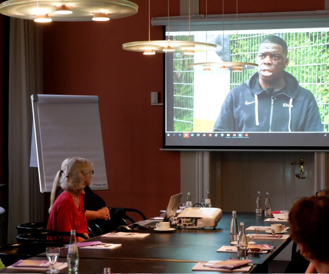 Im Vordergrund ein langer Konferenztisch, an dem Menschen sitzen. Sie schauen in den Bildhintergrund auf eine Leinwand, auf der ein Video zu sehen ist. In dem Video spricht ein Mann wie in einem Interview in die Kamera.