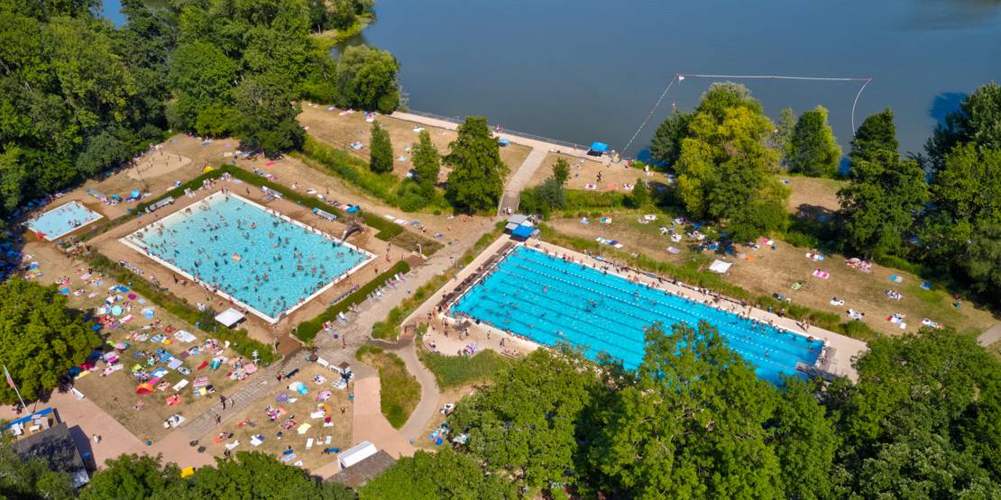 Luftbild eines gut besuchten Schwimmbades mit drei Becken, Liegewiesen und angrenzender Seefläche