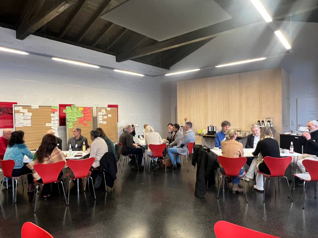 Ein Raum mit mehreren Gruppentischen. Drei der Gruppen sind zu sehen. An den Tischen sitzen jeweils mehrere Menschen unterschiedlichen Alters und Geschlechts. Links im Hintergrund Pinnw&auml;nde.