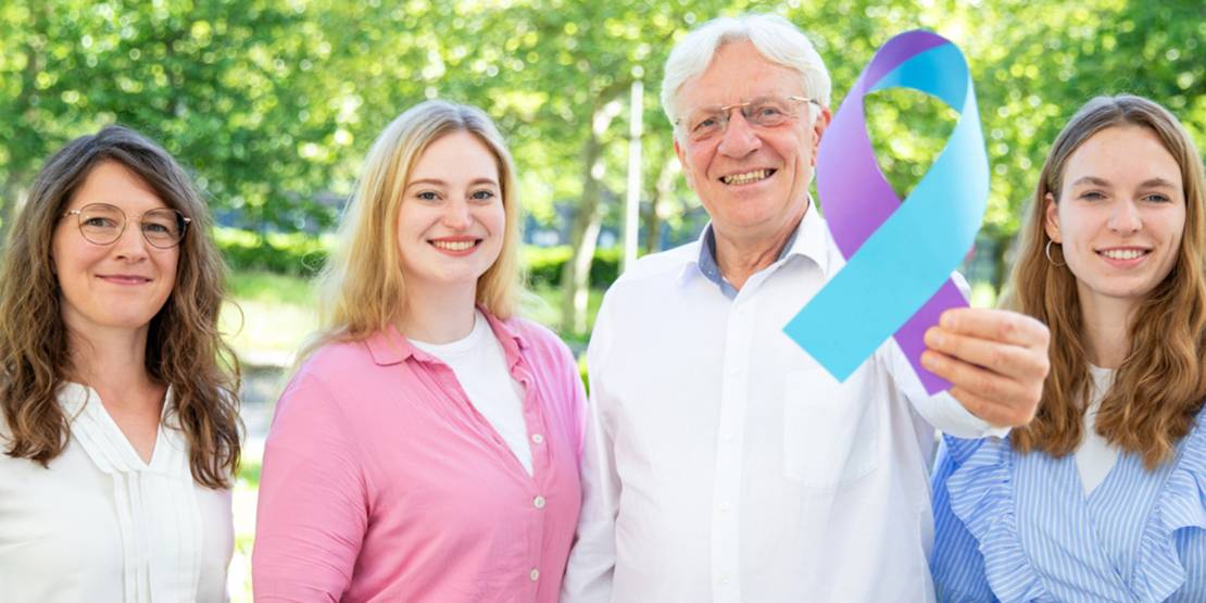 Drei Frauen und ein Mann halten eine blau-lila Schleife in die Kamera.