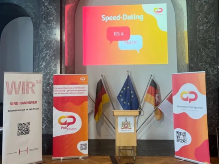 Ein Redepult umgeben von Aufstellern. Ein wei&szlig;er Aufsteller mit dem Text "WIR sind Hannover", zwei orange-roter Aufsteller des Vereins Generation Postmigration. &Uuml;ber dem Redepult ist eine Powerpoint-Folie an die Wnad projeziert mit dem Text "Speed-Dating, It's a match".