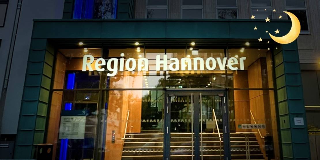 Ein Gebäude mit einem großen Eingang aus Glas der beleuchtet ist, der übrige Teil des Bilds ist dunkel. Über dem Eingang steht "Region Hannover". Oben rechts in der Ecke sind ein Mond und Sterne als grafische Elemente eingefügt.