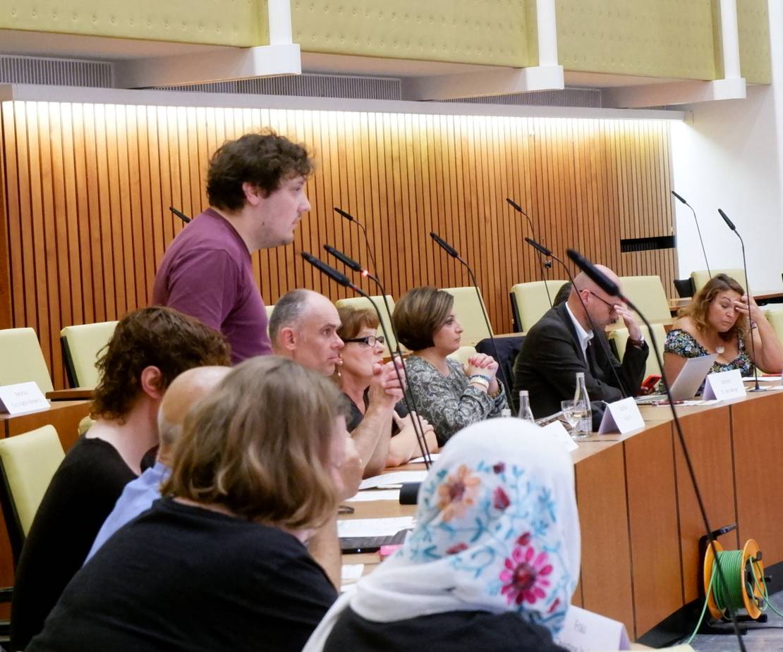 Ratsleute sitzen an den Tischen des Plenums im Ratssaal der Landeshauptstadt Hannover. Einer von ihnen, in der linken Bildhälfte, steht und spricht in ein Tischmikrofon. Er trägt ein violettes Oberteil.