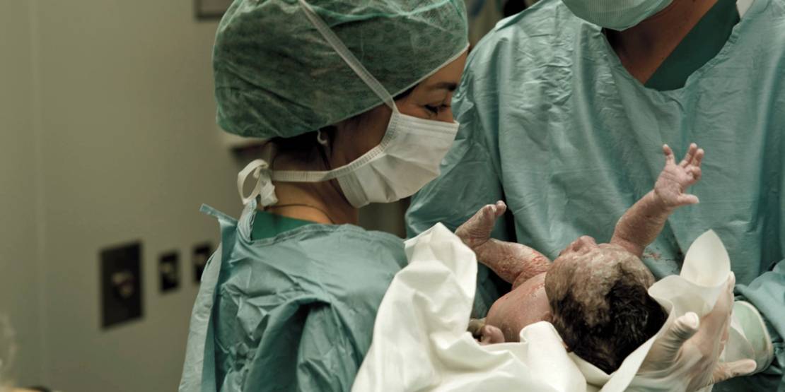 Frau in medizinischer Schutzkleidung hält ein Neugeborenes unmittelbar nach der Geburt in den Händen.