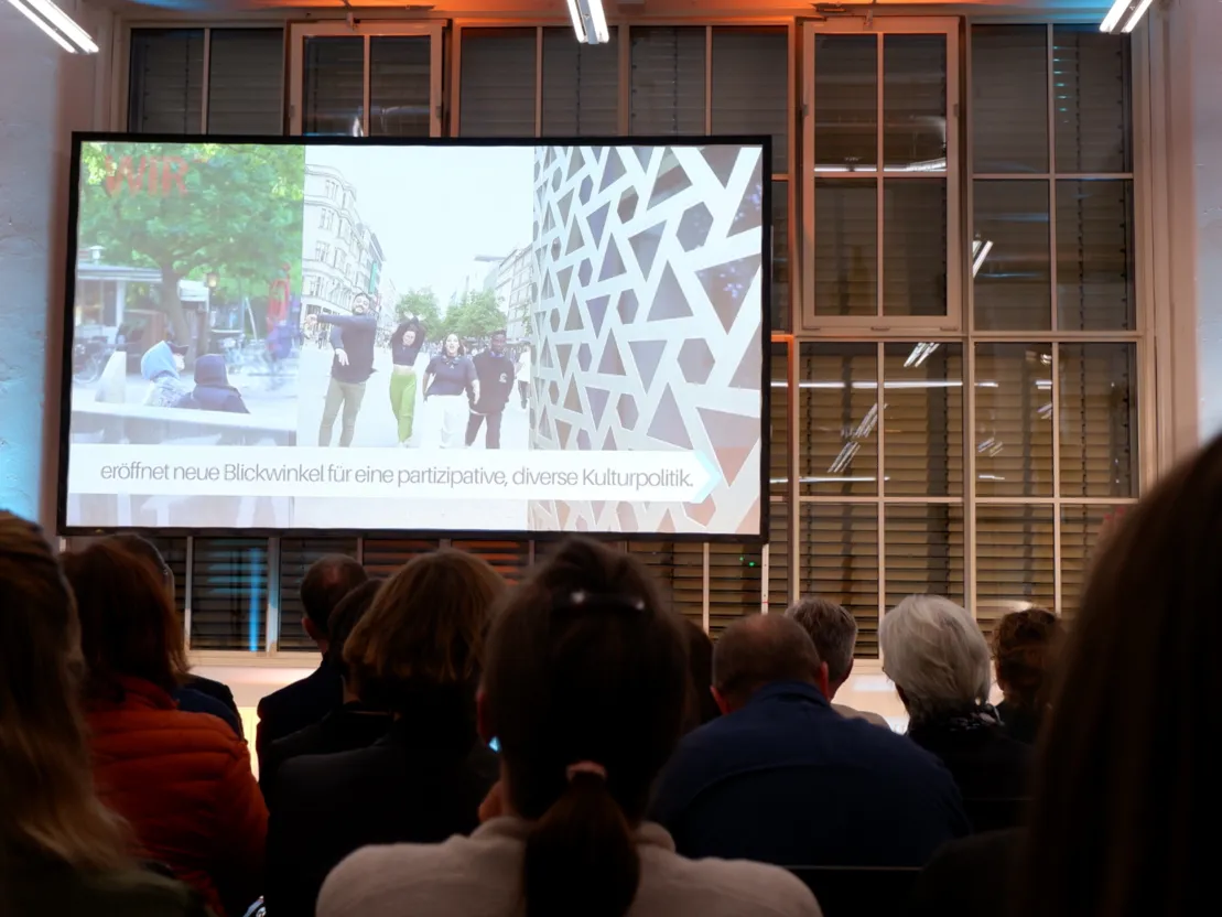 Auf einer Leinwand ein Bild von tanzenden Menschen. Im Vordergrund die hinterk&ouml;pfe eines Publikums, das auf die Leinwand schaut.