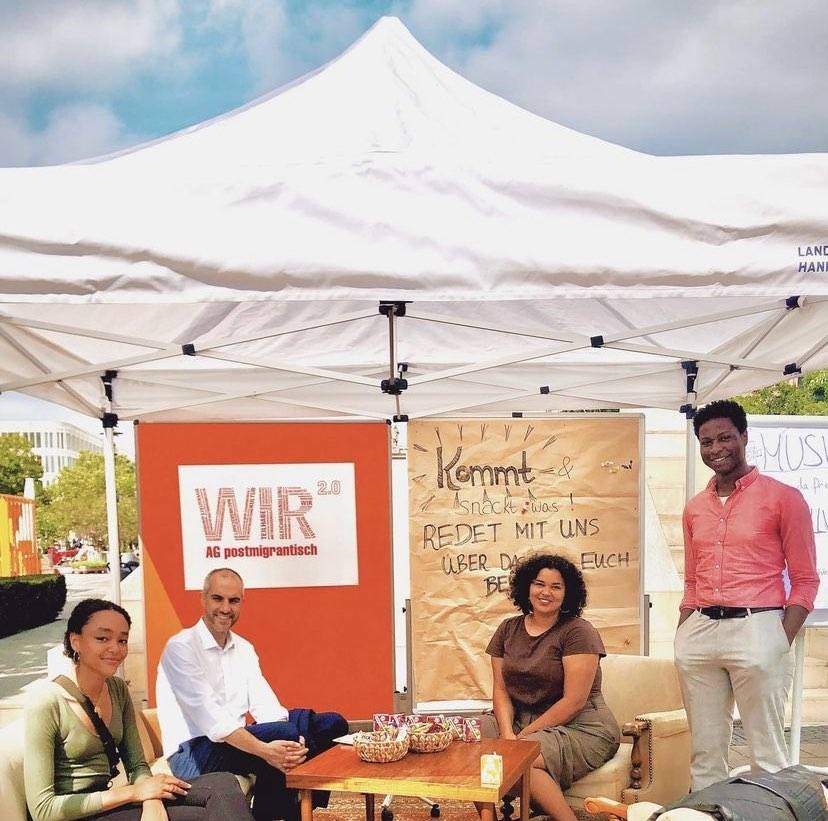 Vier Personen sitzen an einem sonnigen Tag unter einem Pavillon um einen Couchtisch herum. Unter ihnen Oberbürgermeister Belit Onay. Im Hintergrund hängt ein Schild mit der Aufschrift "WIR 2.0 - AG postmigrantisch".