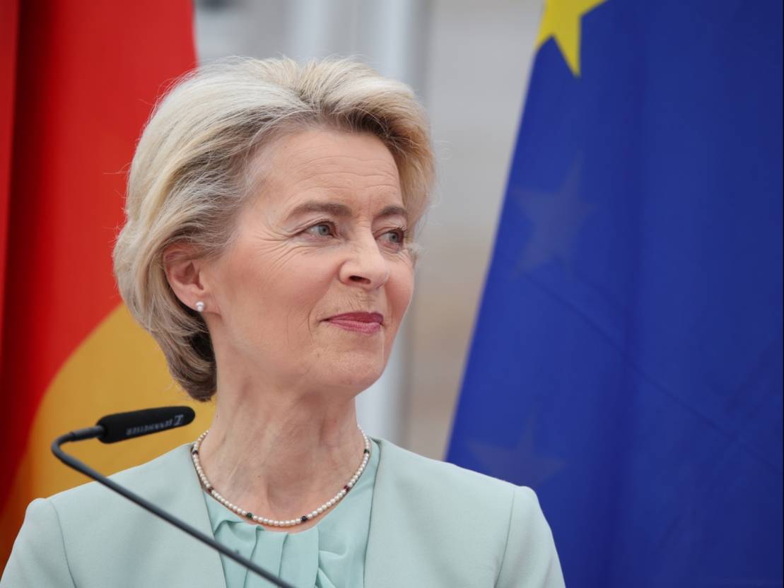 Eine Frau mit blond-grauen Haaren, steht vor zwei Flaggen und schaut aus Betrachtungsperspektive nach rechts. Hinter ihr stehen links die deutsche Flagge und rechts die der Europ&auml;ischen Union.