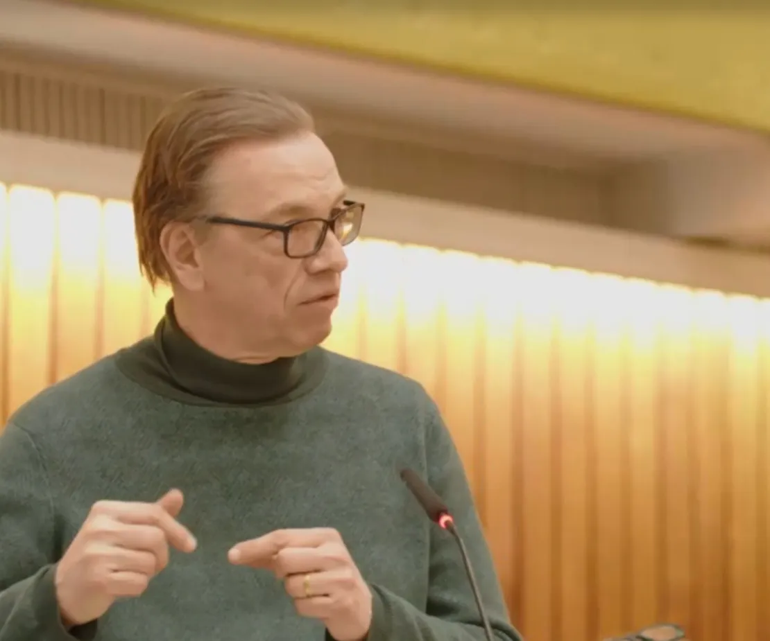 Ein Mann in einem gr&uuml;n-grauen Rollkragenpullover spricht in ein Mikrofon. Er tr&auml;gt eine schwarze Brille und gestikuliert mit beiden H&auml;nden.